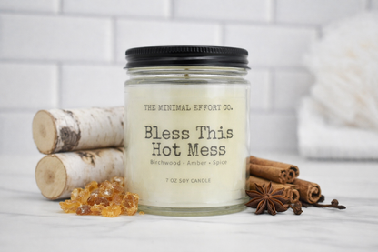 Candle labeled 'Bless This Hot Mess' with cinnamon sticks and star anise on a white surface.