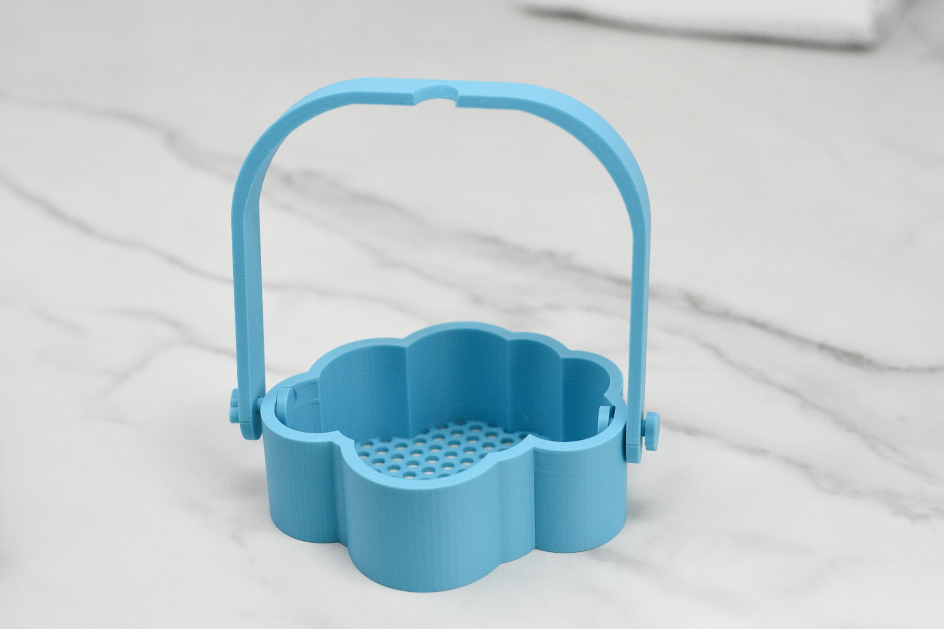 A Bubble Bath Strainer by 570 Soap Co., designed in a blue cloud shape with a handle, sits elegantly on a marble surface, ready to transform your sink into a luxurious oasis. Its small perforations at the bottom guarantee perfect drainage, making it the ideal companion for creating the ultimate bubble bath experience.