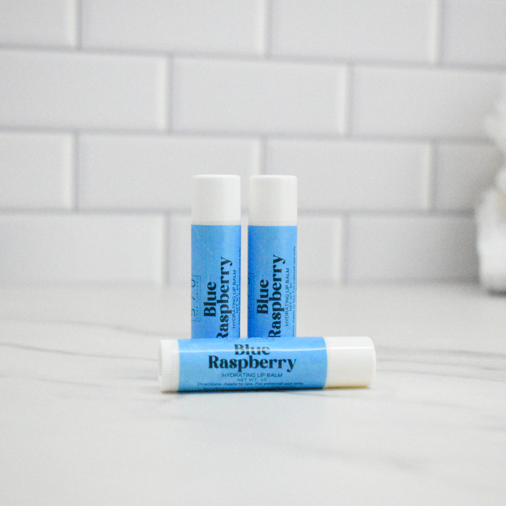 Three lip balm sticks labeled 'Blue Raspberry' on a white surface with a tiled wall background.