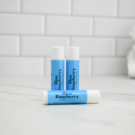 Three lip balm sticks labeled 'Blue Raspberry' on a white surface with a tiled wall background.
