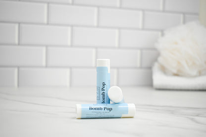 Two lip balm tubes labeled 'bomb Pop' on a bathroom counter with a tiled wall background.