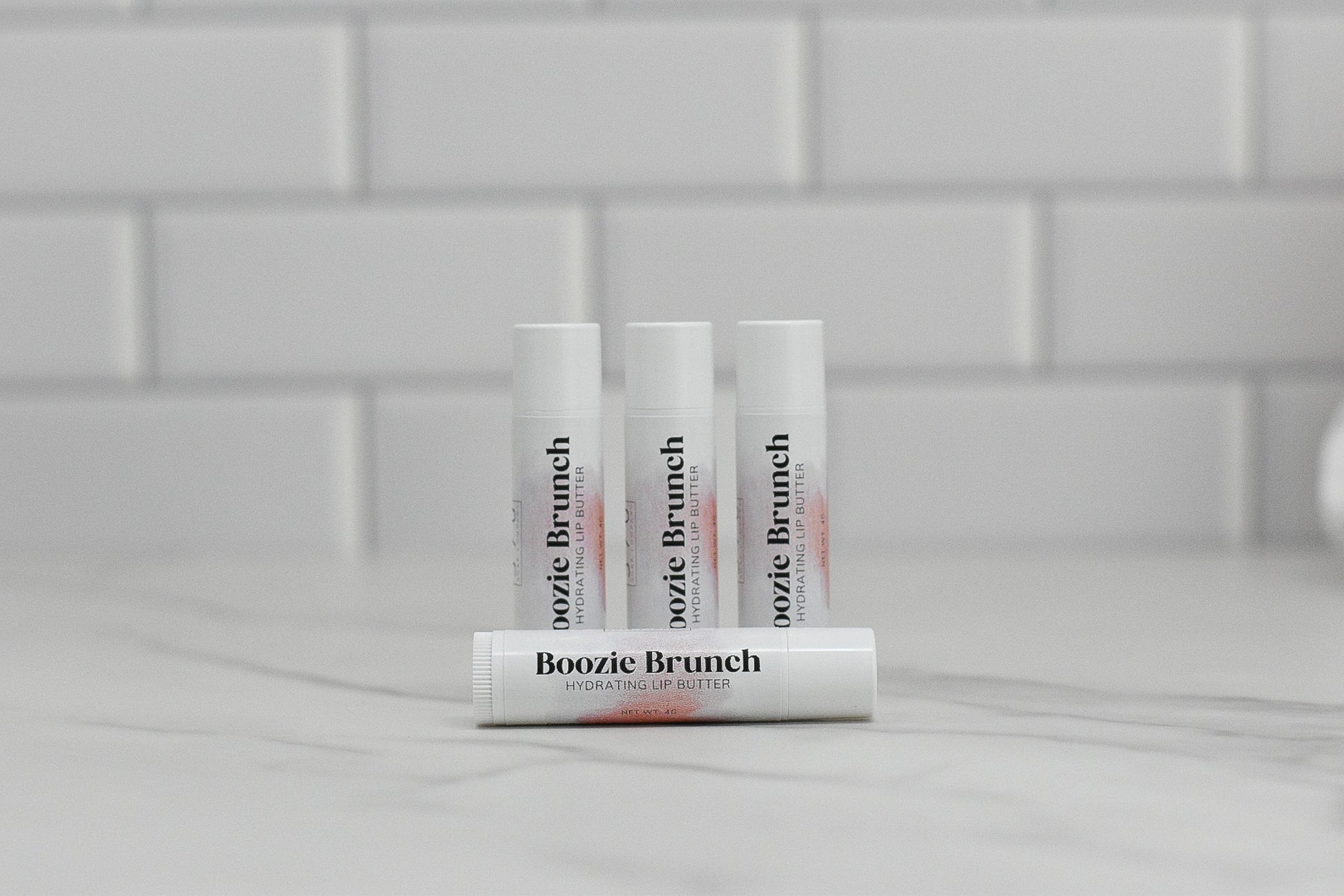 Four tubes of 570 Soap Co.'s Boozie Brunch Peach Bellini Lip Balm rest on a marble surface. White subway tiles in the backdrop contribute to the clean and minimalist atmosphere. Enhance your lip care routine with the luscious scent of Peach Bellini infused in each Boozie Brunch Lip Balm.