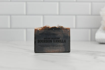 A bar of 570 Soap Co.'s Bourbon Vanilla and Charcoal Bar Soap rests on a marble surface with a white tiled wall in the background. This dark, rustic soap boasts an activated charcoal and skin-nourishing formula. The packaging displays text highlighting its ingredients and benefits.