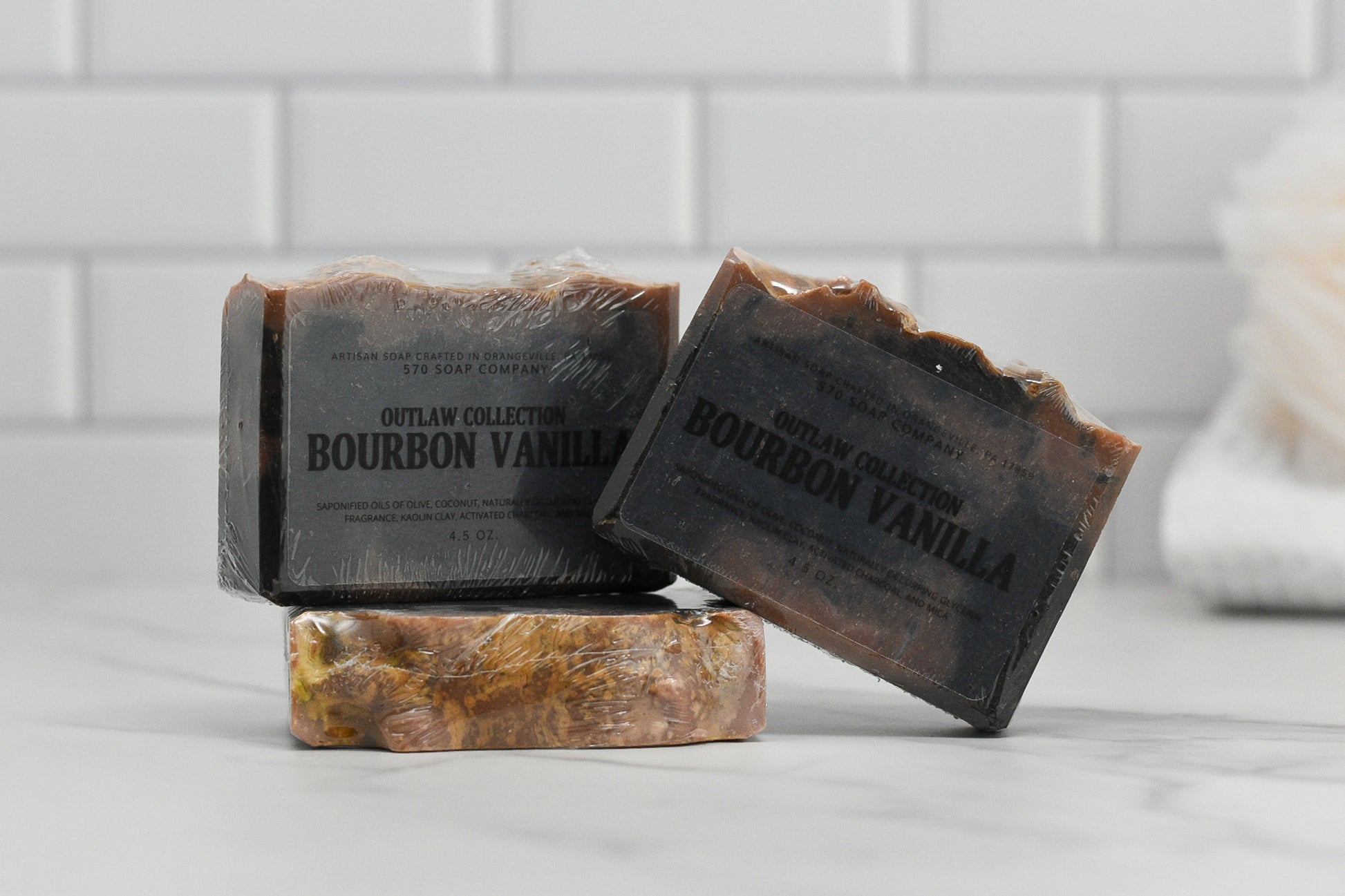 Three bars of Bourbon Vanilla and Charcoal Bar Soap from 570 Soap Co. elegantly rest on a marble surface, with one standing upright, another leaning against it, and the third lying flat. Infused with activated charcoal, this nourishing formula promises a refreshing cleanse against a backdrop of white tiles.