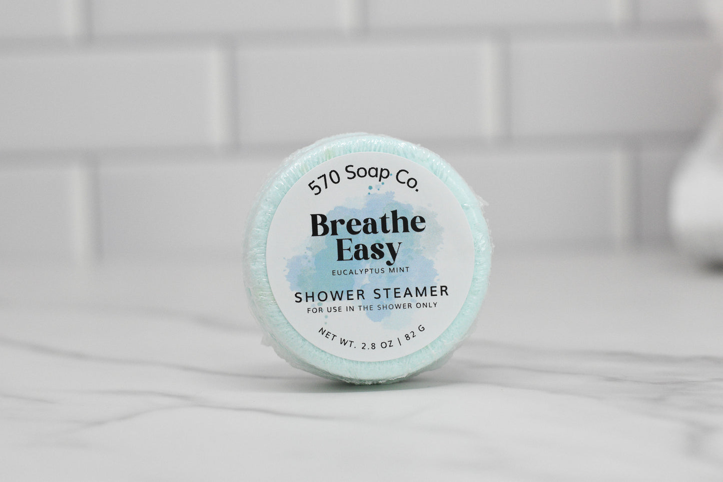 The Breathe Easy Shower Steamer - Eucalyptus Mint by 570 Soap Co. delivers a fragrant experience with its refreshing scent of eucalyptus and mint. The round steamer, set on a white marble surface against a clean tile backdrop, is packaged in light blue with bold black lettering.