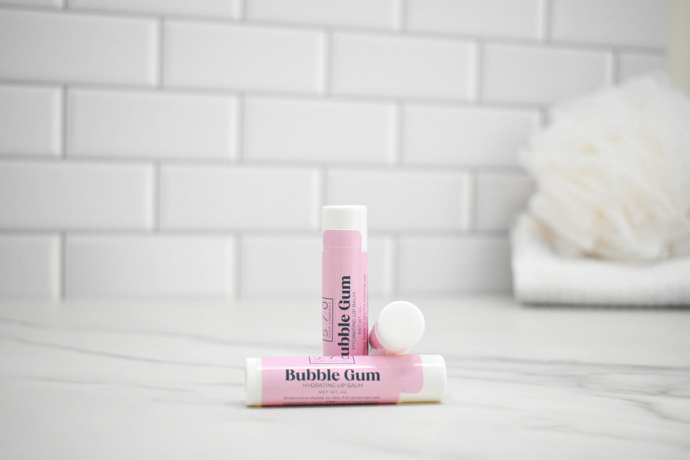 Two lip balm tubes labeled 'Bubble Gum' on a white surface with a tiled wall background.