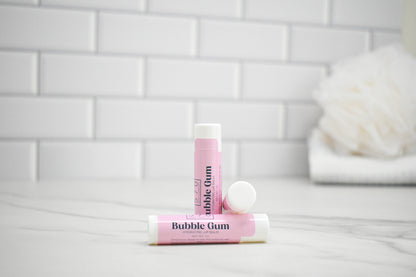 Two lip balm tubes labeled 'Bubble Gum' on a white surface with a tiled wall background.
