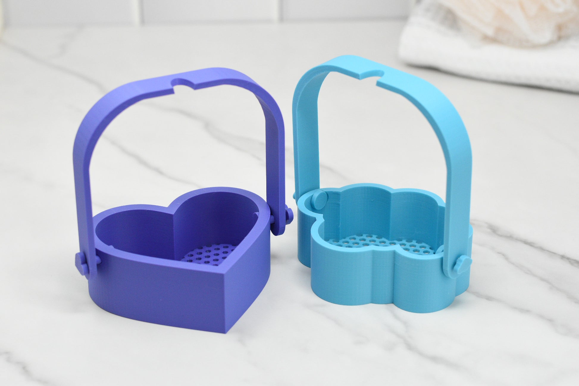 On a marble countertop, two Bubble Bath Strainers from 570 Soap Co. create a luxurious oasis. One strainer is purple with a heart shape, while the other is blue with a flower pattern. Both have convenient handles and drainage holes designed for your next foamy bubble experience. A white towel is in the background, adding to the serene setting.