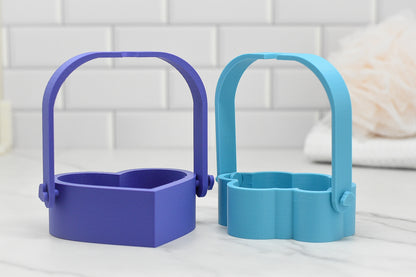 On a marble countertop against a white tile background, two Bubble Bath Strainers by 570 Soap Co. are displayed. The purple strainer is heart-shaped, providing the allure of a luxurious oasis, while the blue one takes the form of a cloud, and both are equipped with handles for convenience.