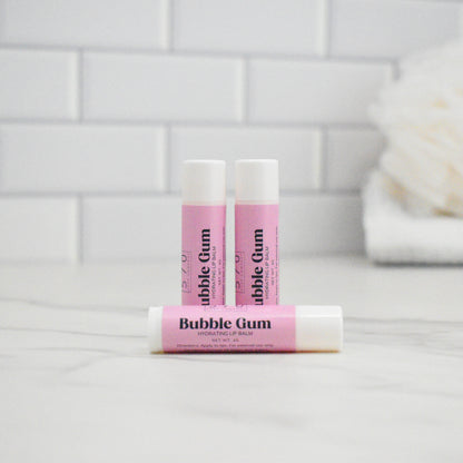 Three lip balms labeled 'Bubble Gum' on a white surface with a tiled wall background.