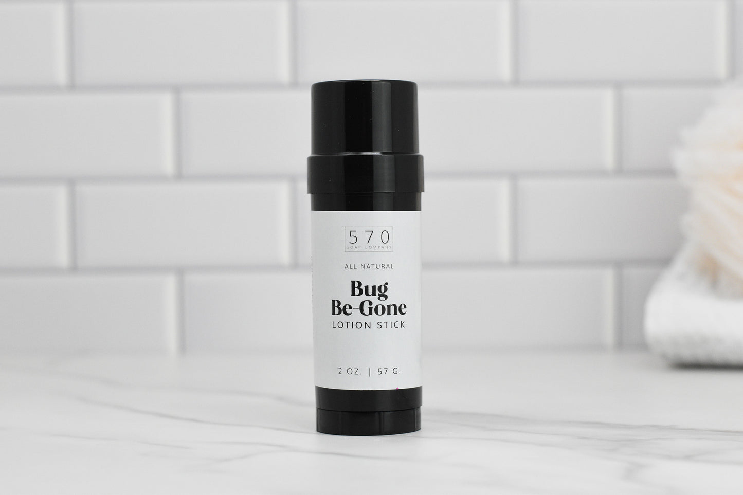 A black and white lotion stick labeled Bug-Be-Gone from 570 Soap Co., crafted with natural ingredients, is positioned on a marble surface. The background showcases white subway tiles.