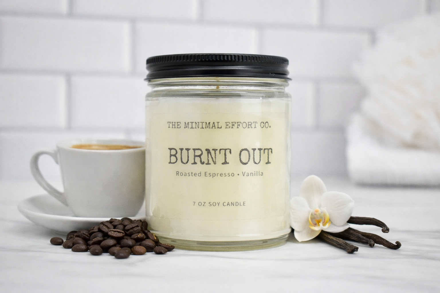 Candle labeled 'Burnt Out' from The Minimal Effort Co. with coffee beans, a cup of coffee, and an orchid on a white surface.