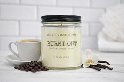 Candle labeled 'Burnt Out' from The Minimal Effort Co. with coffee beans, a cup of coffee, and an orchid on a white surface.