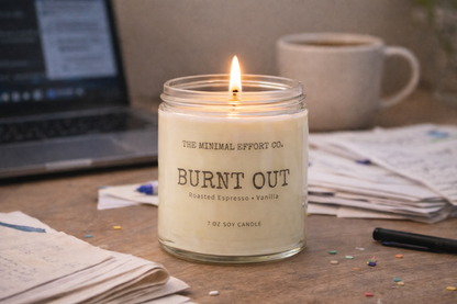 Candle labeled 'Burnt Out' from The Minimal Effort Co. on a desk with a laptop and mug in the background.