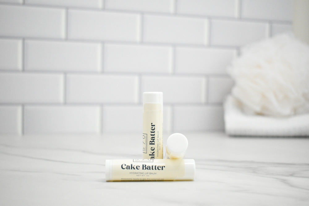 Two lip balm tubes labeled 'Cake Batter' on a white surface with a tiled wall background.