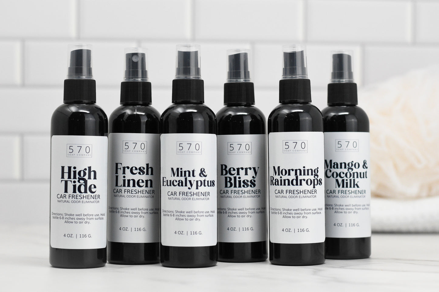 Six bottles of Linen & Room Spray from 570 Soap Co., each offering a unique fragrance, are lined up on a white countertop. Scents include High Tide, Fresh Linen, and Mint & Eucalyptus. These natural odor eliminators feature simple monochrome labels and capture the essence of refreshing car fresheners.