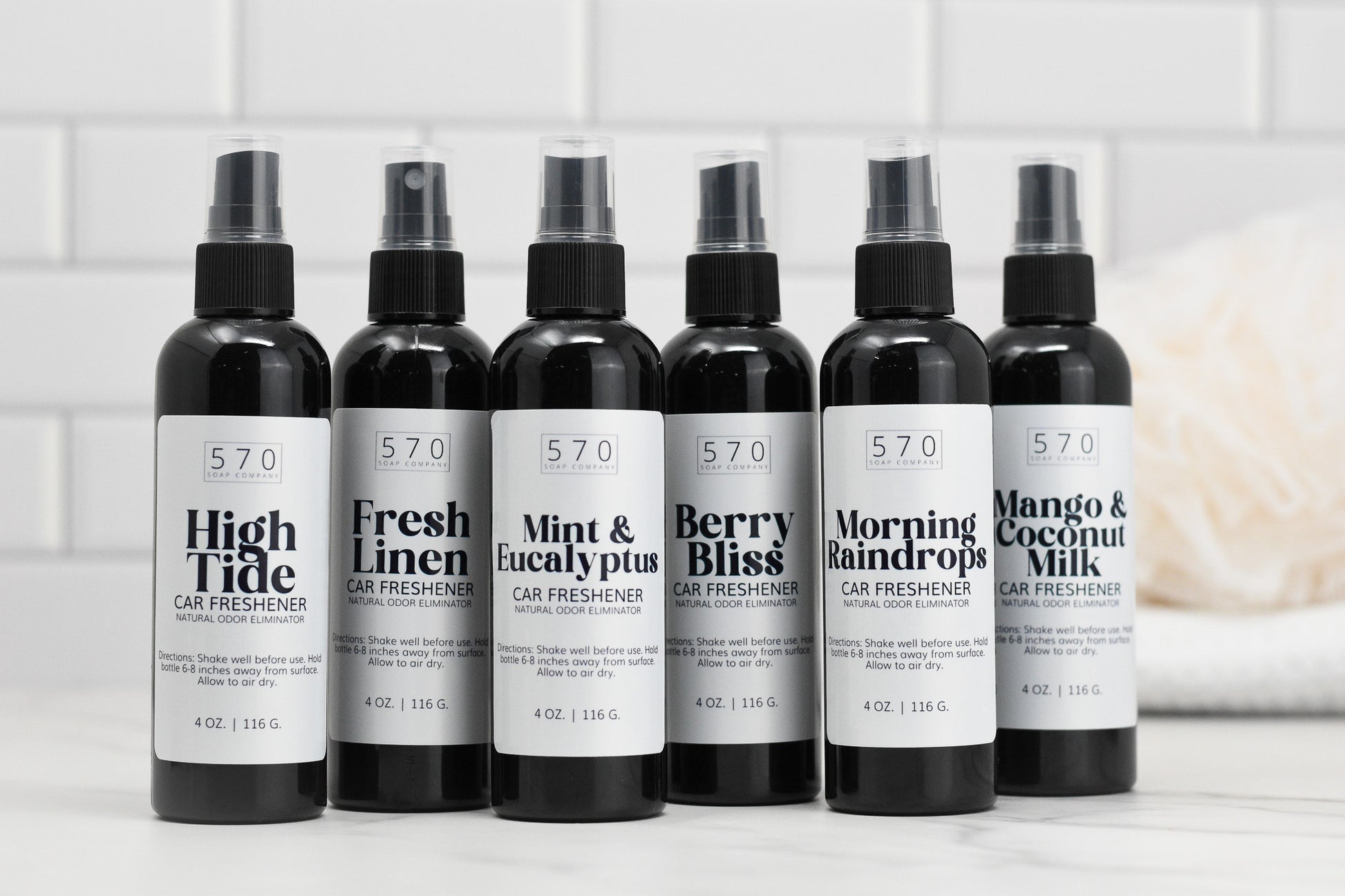 Six bottles of Linen & Room Spray from 570 Soap Co., each offering a unique fragrance, are lined up on a white countertop. Scents include High Tide, Fresh Linen, and Mint & Eucalyptus. These natural odor eliminators feature simple monochrome labels and capture the essence of refreshing car fresheners.