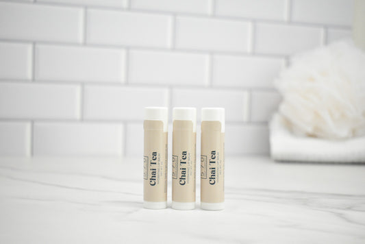 Three lip balm tubes with 'Chai Tea' branding on a white surface with a tiled wall background.