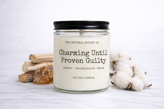 Candle jar with label 'Charming Until Proven Guilty' on a white background with cotton and wood elements.