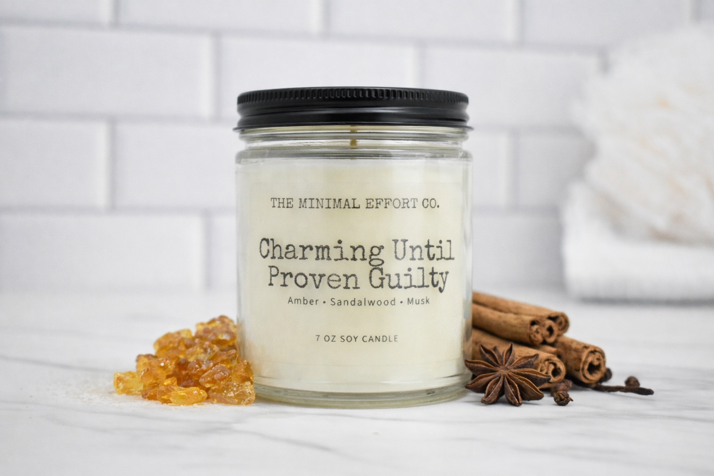 Candle jar labeled 'Charming Until Proven Guilty' by The Minimal Effort Co. on a white surface with cinnamon sticks and star anise.