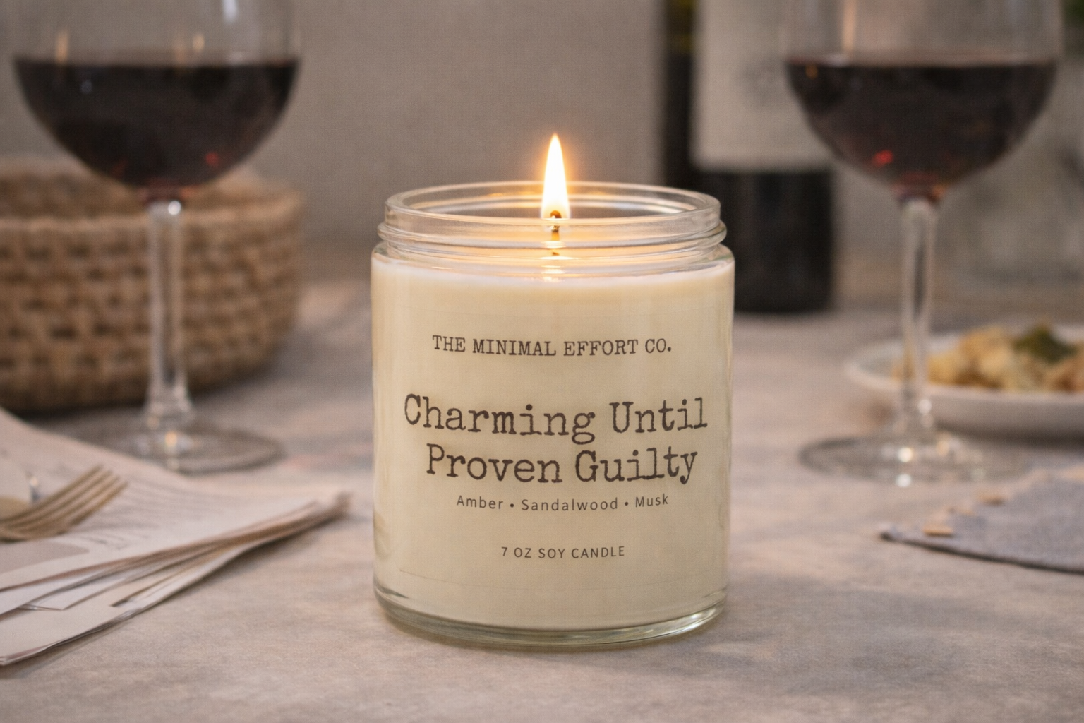 Candle labeled 'Charming Until Proven Guilty' on a table with wine glasses and food.