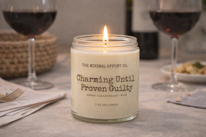 Candle labeled 'Charming Until Proven Guilty' on a table with wine glasses and food.