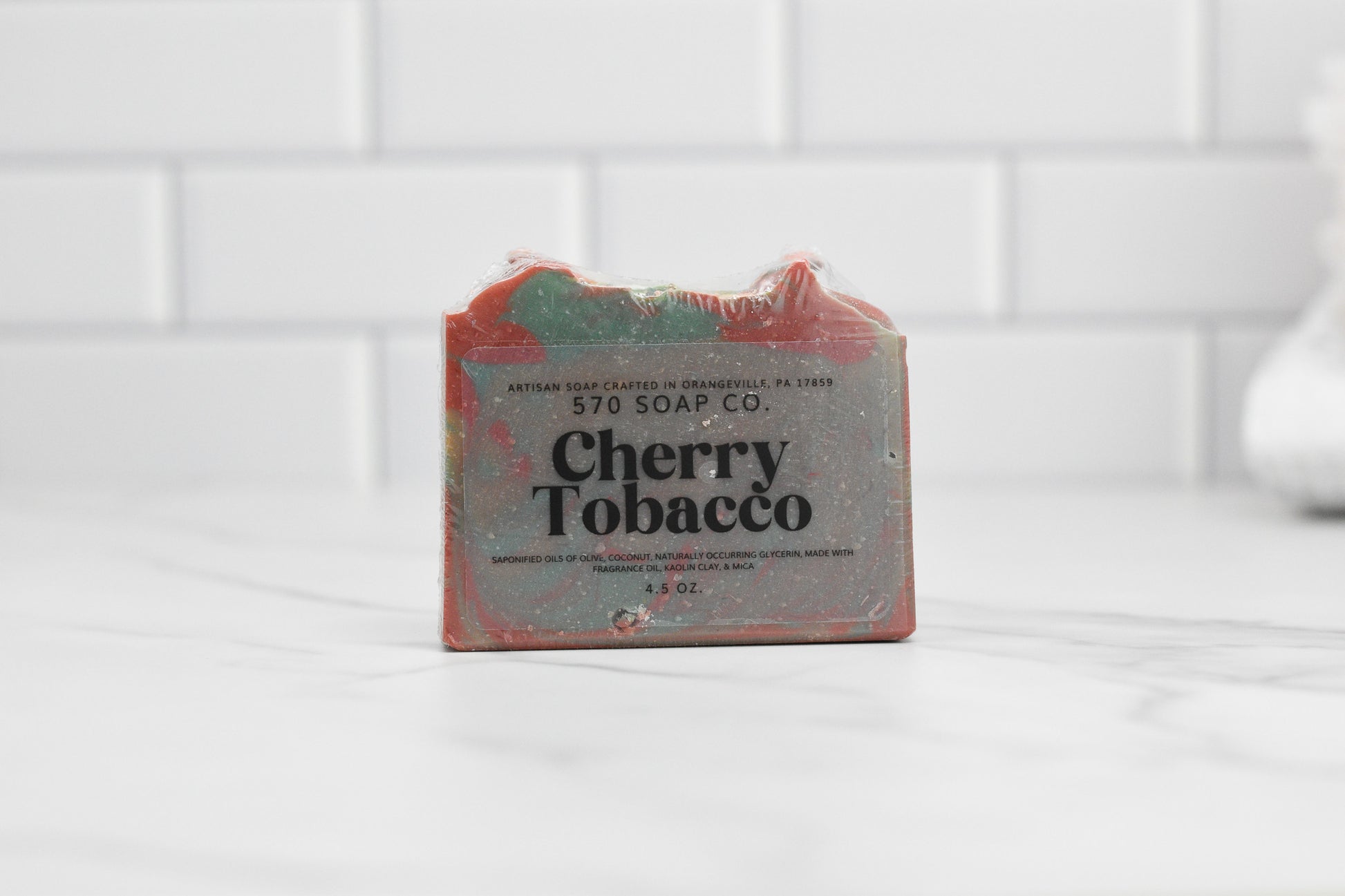 The Cherry Tobacco Bar Soap by 570 Soap Co. rests elegantly on a marble surface with a backdrop of white tiles. This handmade soap, infused with the essence of cherrywood and kaolin clay, showcases striking red and green accents that enhance any bath space with its rustic allure.