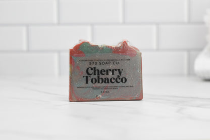 The Cherry Tobacco Bar Soap by 570 Soap Co. rests elegantly on a marble surface with a backdrop of white tiles. This handmade soap, infused with the essence of cherrywood and kaolin clay, showcases striking red and green accents that enhance any bath space with its rustic allure.