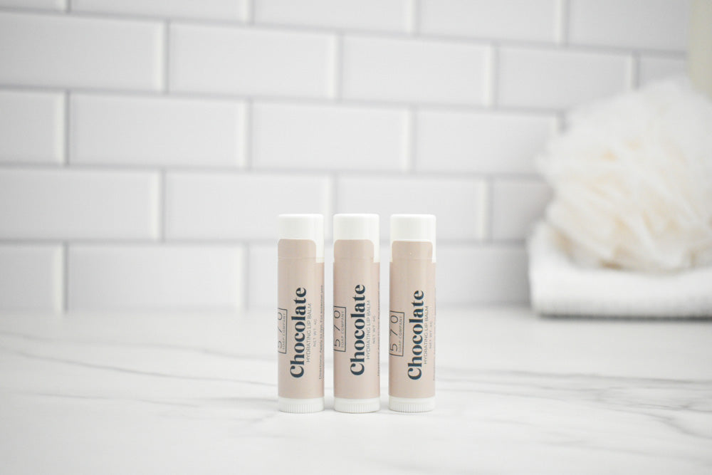 Three lip balm tubes with 'Chocolate' branding on a white surface against a tiled wall background.