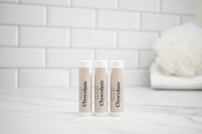 Three lip balm tubes with 'Chocolate' branding on a white surface against a tiled wall background.