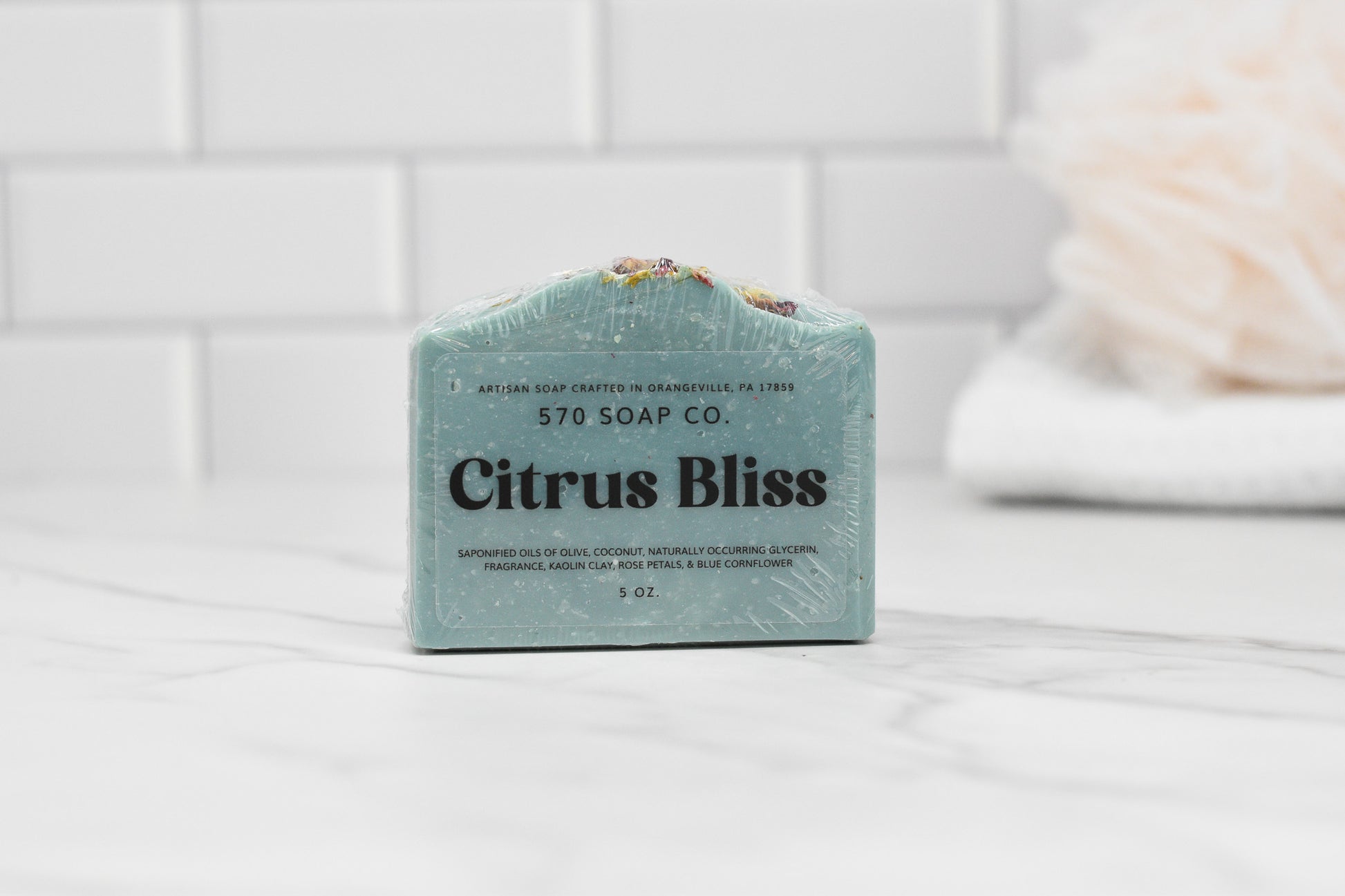 A Citrus Bliss Soap Bar by 570 Soap Co. sits on a marble surface, releasing an invigorating citrus aroma. The transparent packaging showcases the green soap as a white towel fades into the background against a tiled wall, offering your skin an indulgent moisturizing experience.