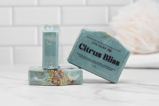 A set of three Citrus Bliss Soap Bars from 570 Soap Co. in a teal hue are artfully arranged on a white marble surface, releasing an invigorating citrus aroma. Two bars are stacked, with one positioned flat and the other propped against it. A white tiled wall serves as the backdrop, accented by a bathroom sponge.