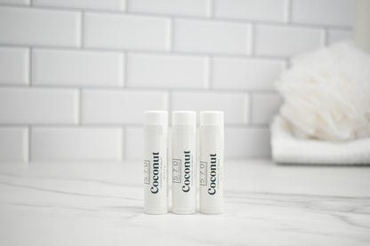 Three lip balm tubes labeled 'Coconut' on a white surface with a tiled wall background.