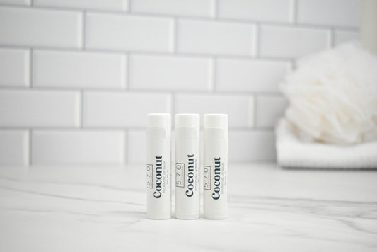 Three lip balm tubes labeled 'Coconut' on a white surface with a tiled wall background.