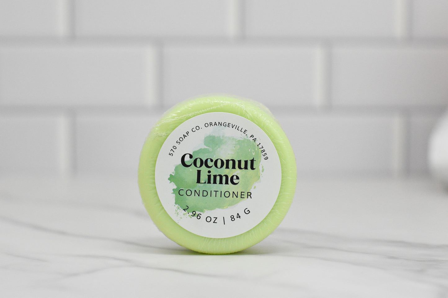 A round bar of 570 Soap Co.'s Conditioner Bar for All Hair Types - Coconut Lime, infused with Tropical Hydration, rests on a marble surface. It is wrapped in light green packaging that displays product details and weighs 3 oz (84 g), harmonizing with the serene white-tiled wall in the background.