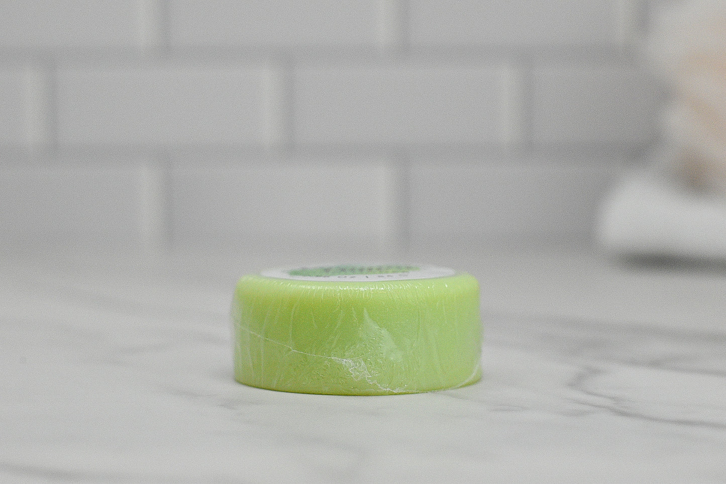 A small, round bar of light green Conditioner Bar for All Hair Types - Coconut Lime from 570 Soap Co. rests on a marble countertop, promising tropical hydration. Wrapped in clear plastic, its refreshing aroma complements the serene backdrop of a white tiled wall.