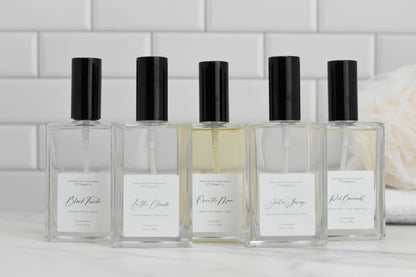 Five rectangular perfume bottles with black caps are lined up against a white tiled background, each prominently displaying a luxury fragrance name. On the right, a beige loofah is partly visible, accentuating the elegance of these Eau de Parfum selections from 570 Soap Co., including the "Over the Moon" fragrance.