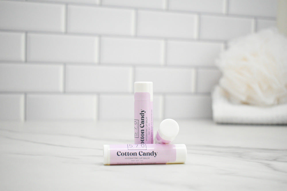Two lip balms labeled 'Cotton Candy' on a white surface with a tiled wall background.