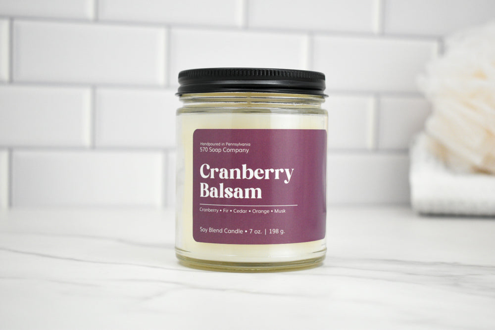 Candle jar labeled 'Cranberry Balsam' on a white surface with a tiled wall background