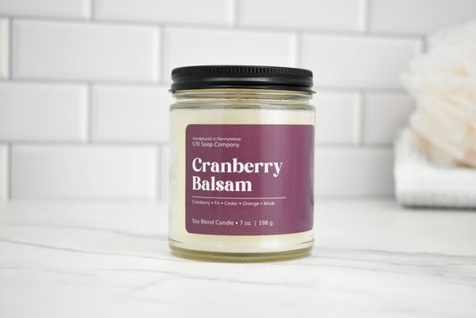 Candle jar labeled 'Cranberry Balsam' on a white surface with a tiled wall background
