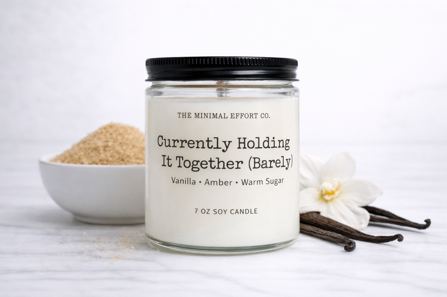 Candle labeled 'Currently Holding It Together (Barely)' with vanilla beans and a flower on a white background