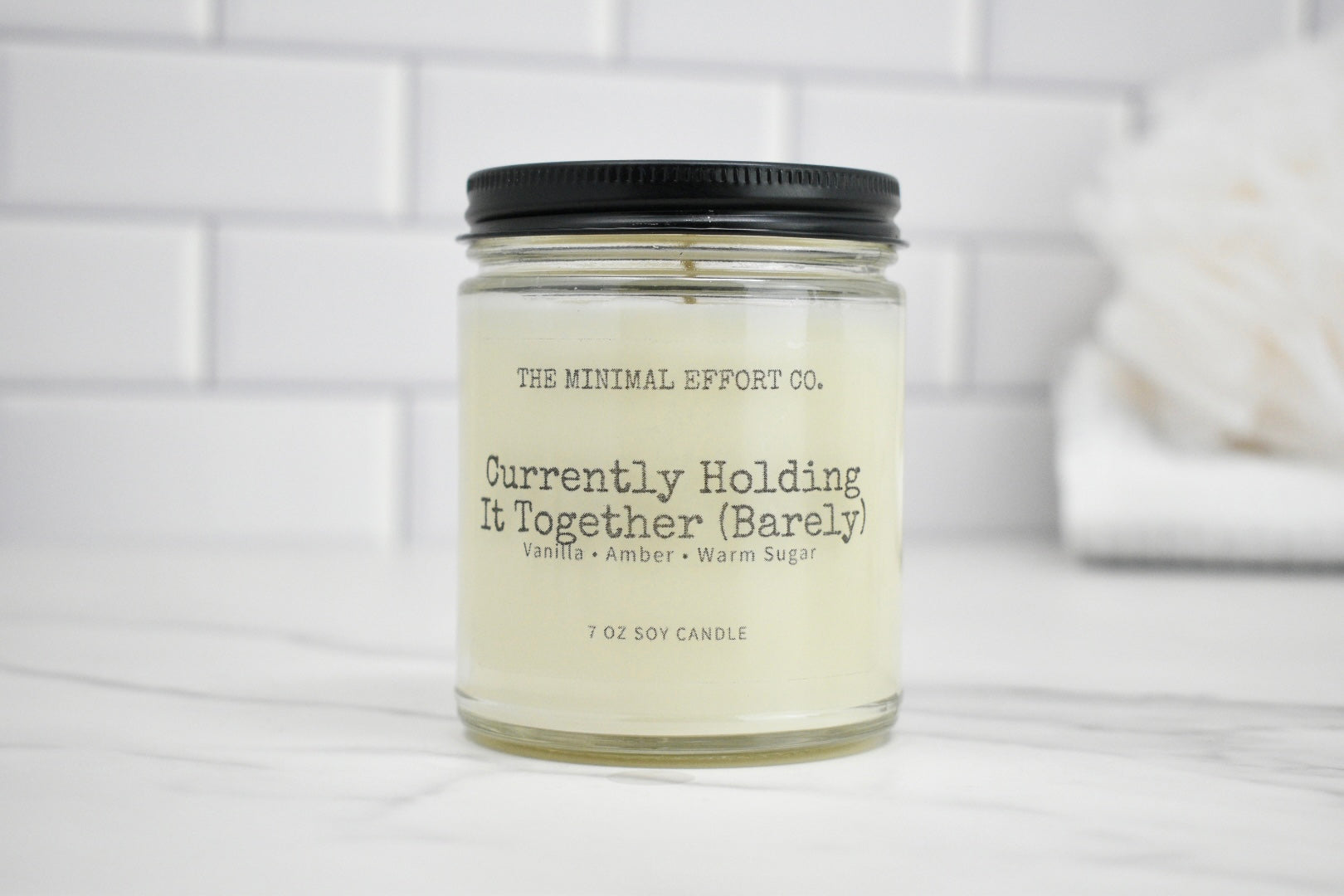 Candle jar labeled 'Currently Holding It Together (Barely)' by The Minimal Effort Co. on a white surface with a white tiled wall background.