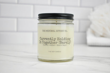 Candle jar labeled 'Currently Holding It Together (Barely)' by The Minimal Effort Co. on a white surface with a white tiled wall background.
