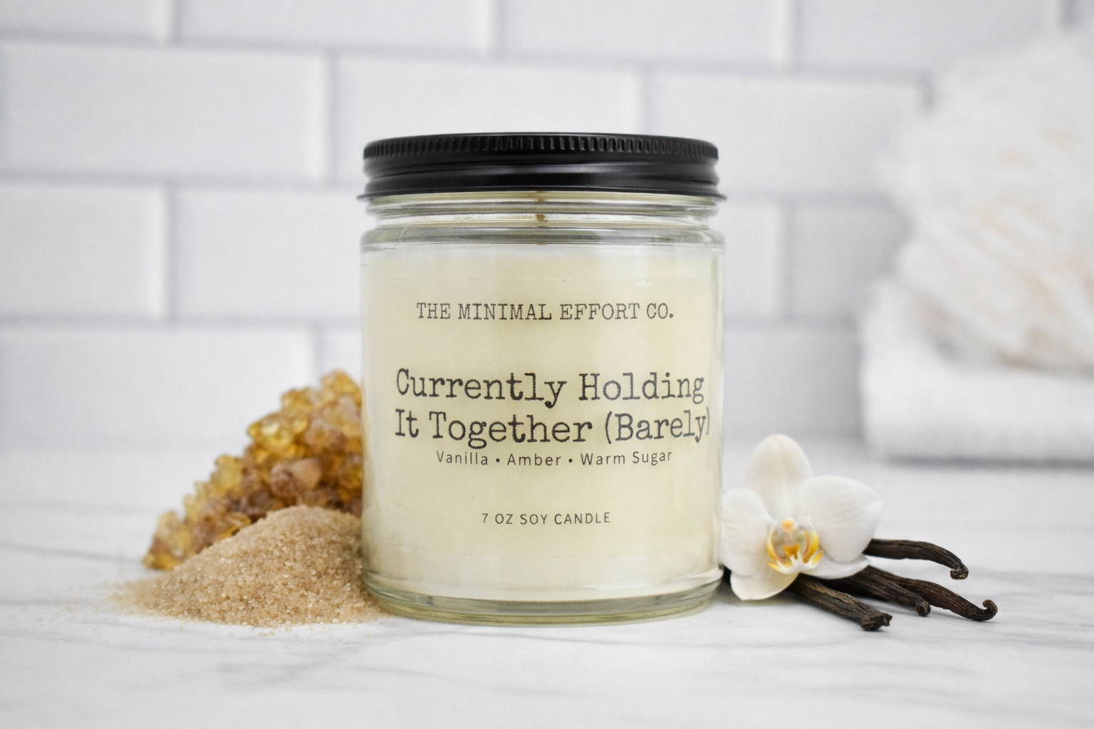 Candle jar labeled 'Currently Holding It Together (Barely)' by The Minimal Effort Co. on a white surface with vanilla beans and an orchid.