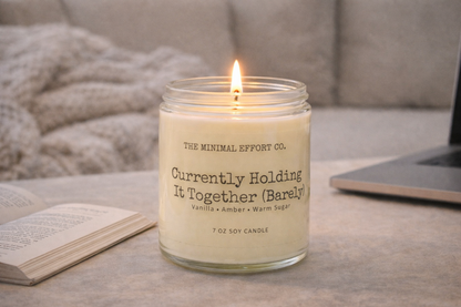 Candle labeled 'Currently Holding It Together (Barely)' by The Minimal Effort Co. on a table with a book and laptop.
