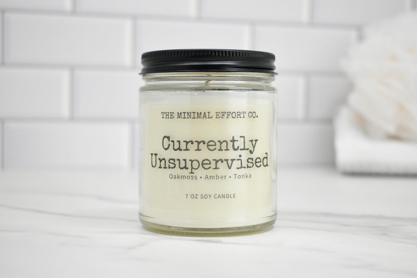 Candle jar labeled 'Currently Unsupervised' by The Minimal Effort Co. on a white surface with a tiled wall background.
