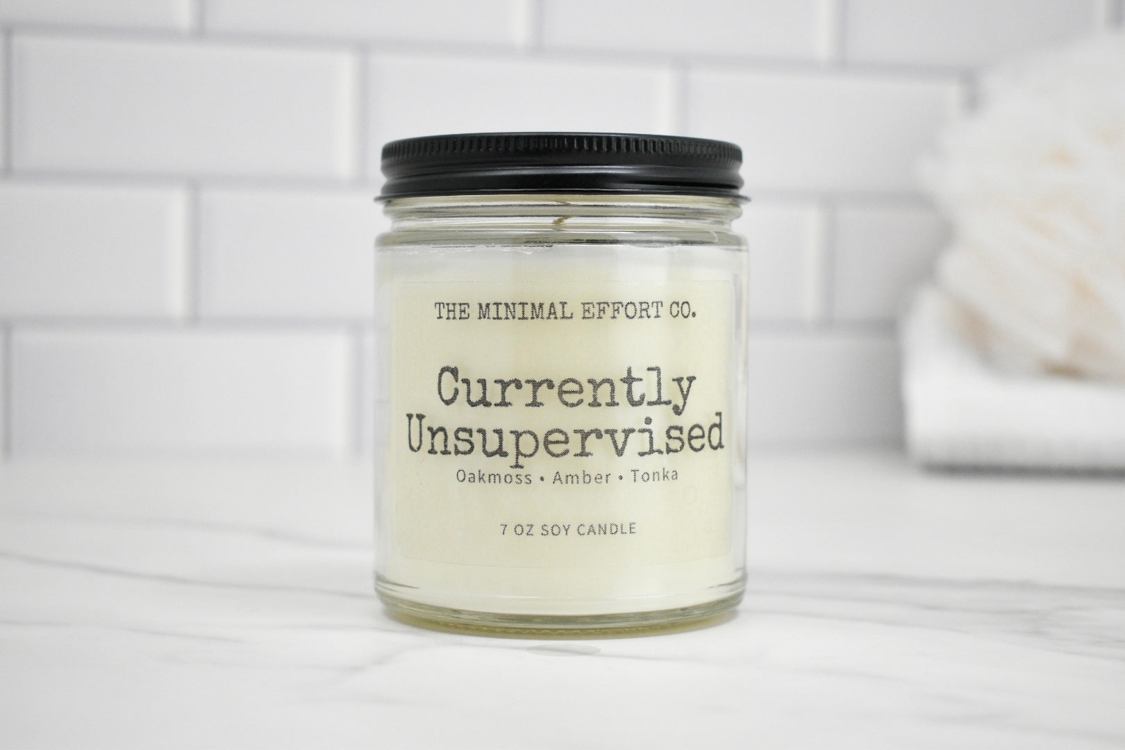 Candle jar labeled 'Currently Unsupervised' by The Minimal Effort Co. on a white surface with a tiled wall background.