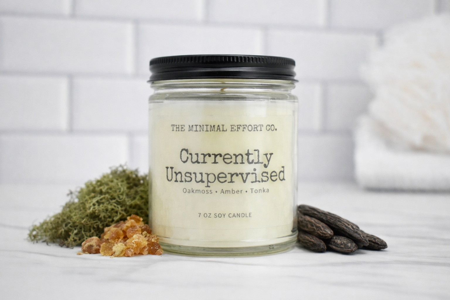 Candle jar labeled 'Currently Unsupervised' from The Minimal Effort Co. on a white surface with decorative elements.