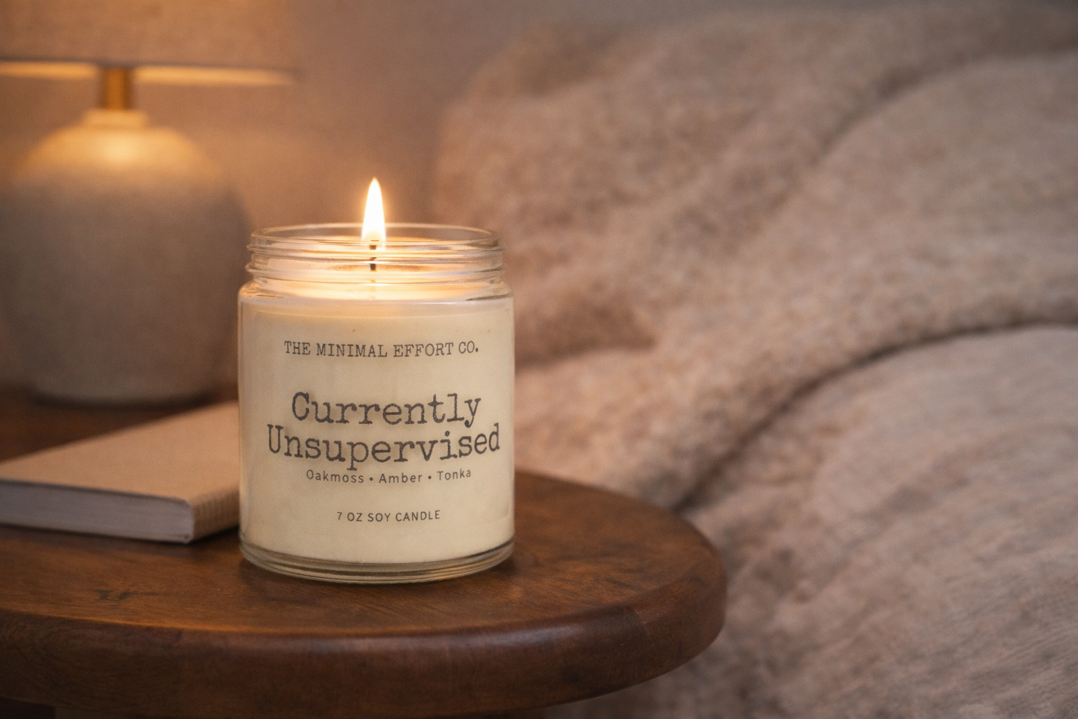 Candle with a label on a wooden table next to a book and lamp, with a blurred background