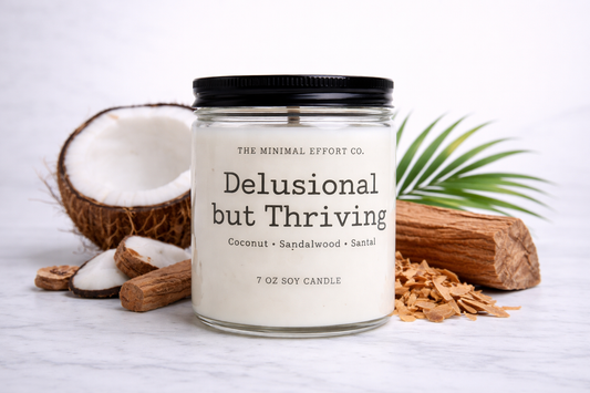 Candle jar labeled 'Delusional but Thriving' with coconut and sandalwood on a light background